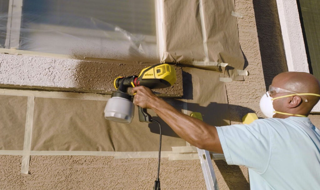 man spraying exterior trim with a paint sprayer while wearing a mask