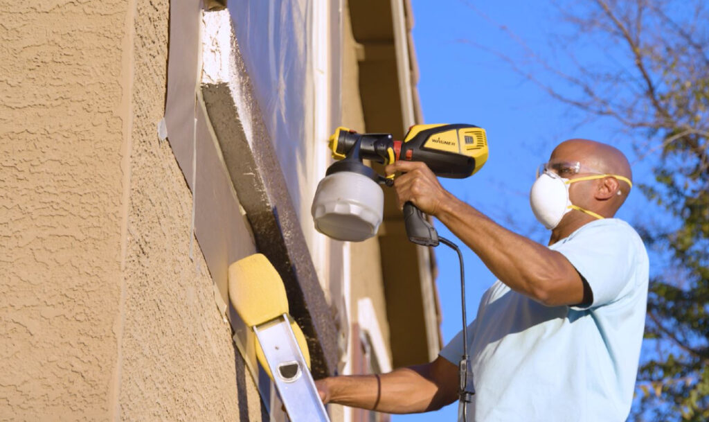 man on ladder spraying exterior trim with a sprayer while wearing mask